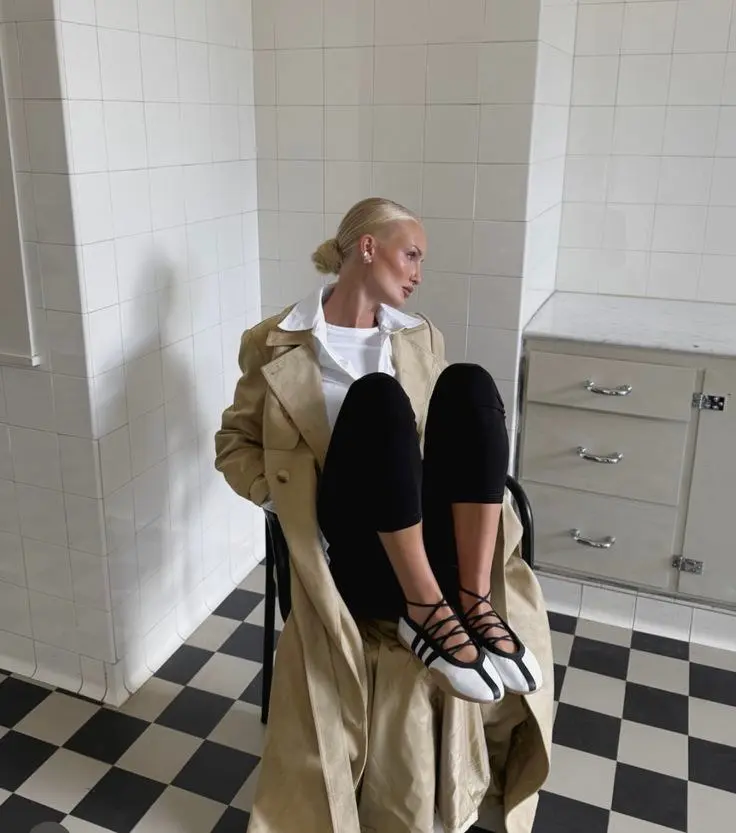 woman seated in chair in camel trench coat white button-up shirt black leggings black and white lace-up ballet sneakers sleek bun pearl earrings tiled interior photo 13