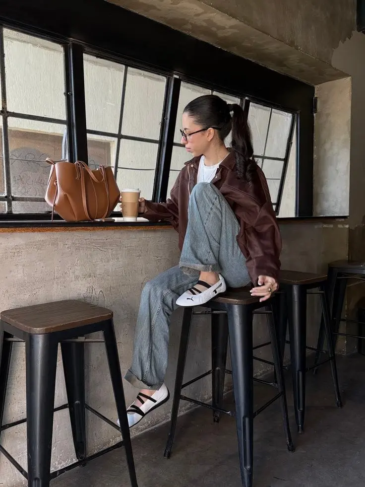 woman seated at coffee shop counter in chocolate brown leather jacket white tee light wash straight-leg jeans white Mary Jane ballet sneakers caramel leather tote bag on counter photo 15