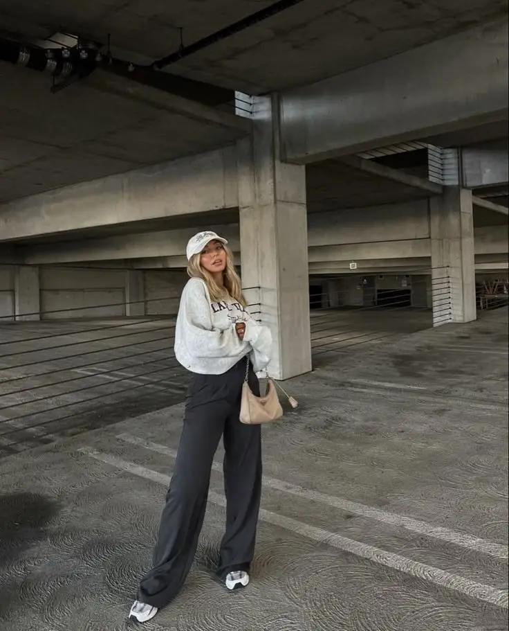 blonde woman wearing grey graphic sweatshirt baseball cap wide leg dark charcoal trousers New Balance white sneakers parking garage outfit 2