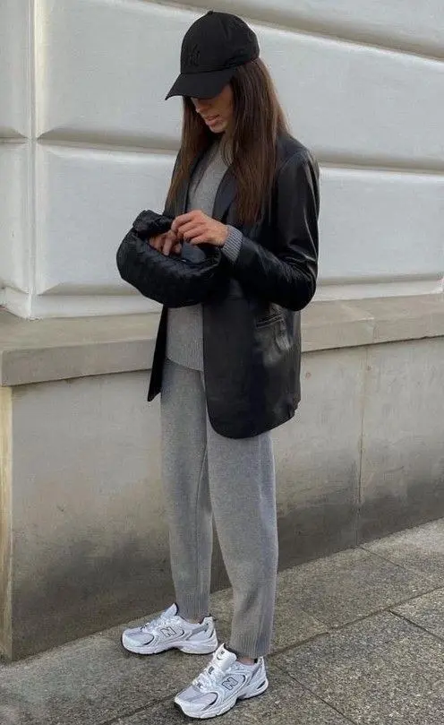 brunette woman wearing black leather blazer grey ribbed knit sweater grey wide leg knit pants black baseball cap woven clutch New Balance white silver sneakers outfit 5