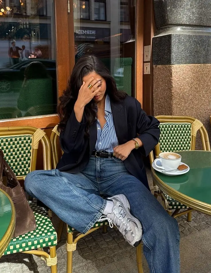 dark-haired woman sitting at café wearing navy pinstripe oversized blazer blue striped shirt medium wash baggy jeans New Balance 530 grey silver sneakers gold ring watch outfit 6