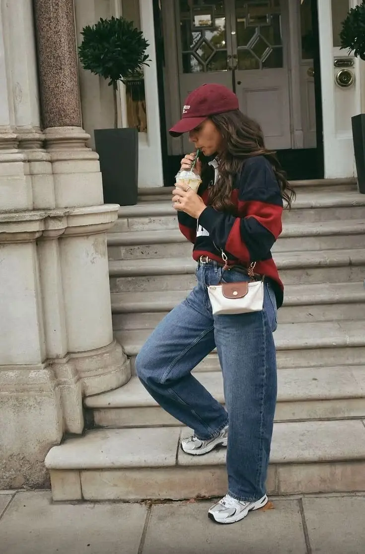brunette woman on steps wearing burgundy cap color-block navy red varsity sweatshirt baggy medium-wash jeans mini nylon belt bag New Balance 530 silver sneakers outfit 7