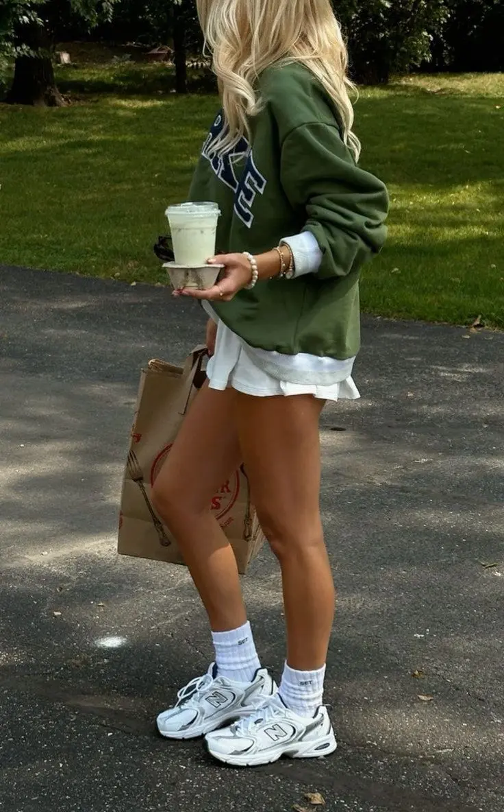 blonde woman from behind wearing oversized green varsity graphic sweatshirt white mini skirt white crew socks New Balance 530 white sneakers pearl bracelet tote bag outfit 9