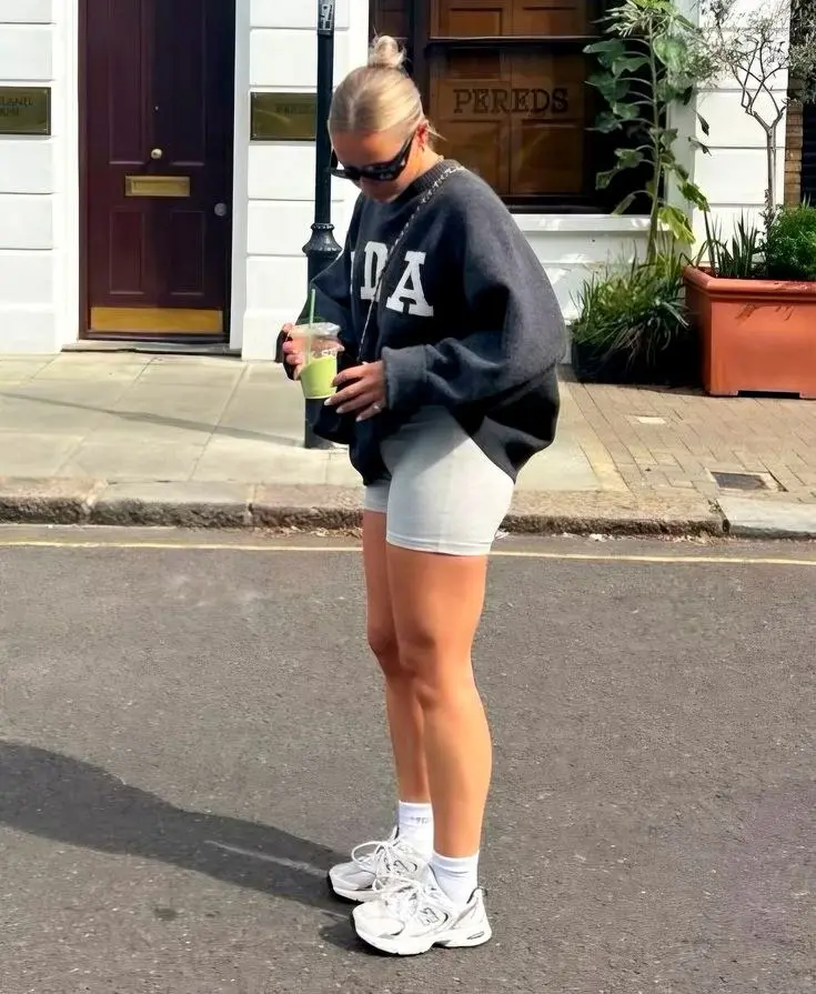 blonde woman wearing charcoal oversized varsity sweatshirt light grey biker shorts white crew socks New Balance 530 white silver sneakers chain bag wraparound sunglasses outfit 13