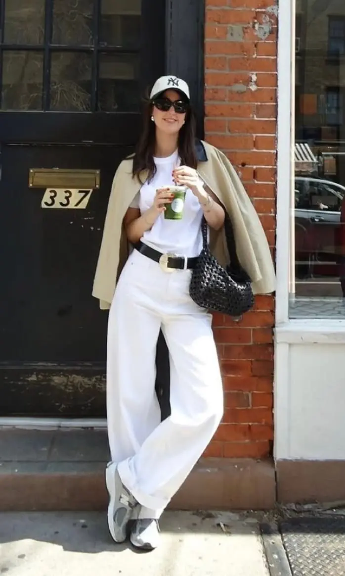 brunette woman wearing white baseball cap white tee white wide-leg jeans black wide belt beige trench coat draped over shoulders New Balance 530 grey silver sneakers black woven tote bag outfit 15
