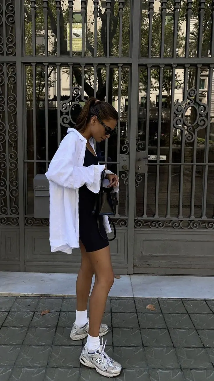 brunette woman wearing oversized white linen shirt open over black mini dress white crew socks New Balance 530 silver grey sneakers small black bucket bag ponytail sunglasses outfit 16