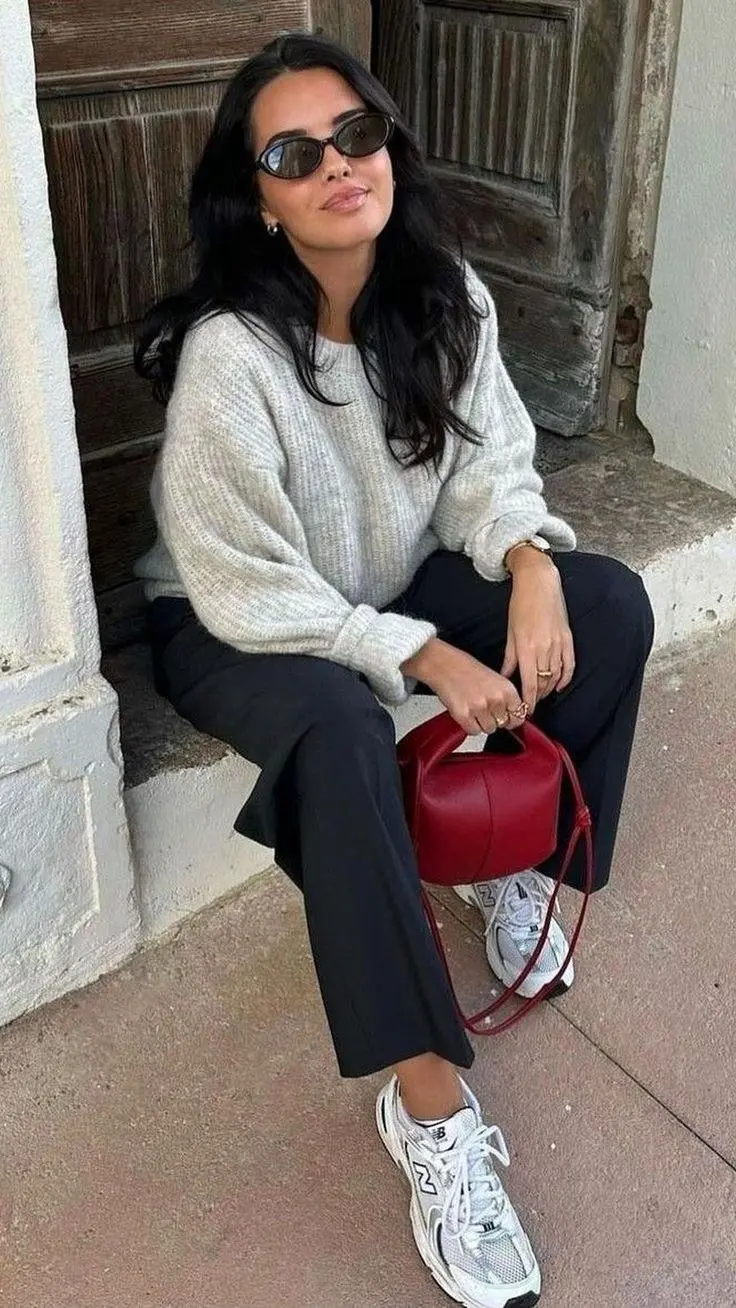 dark-haired woman sitting wearing oval sunglasses oat light grey chunky knit sweater black wide leg tailored trousers New Balance 530 white silver sneakers red structured mini bucket bag outfit 17
