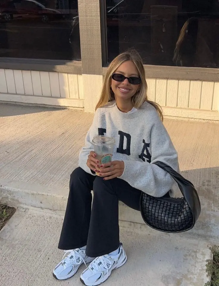 blonde woman sitting on sidewalk wearing oat grey varsity letter graphic knit sweater black flared wide leg trousers New Balance 530 white silver sneakers black woven hobo bag round sunglasses outfit 18