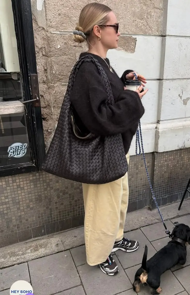 woman in dark brown oversized knit sweater with large woven dark brown shoulder bag cream wide leg trousers and chunky black athletic sneakers standing on sidewalk outfit 1
