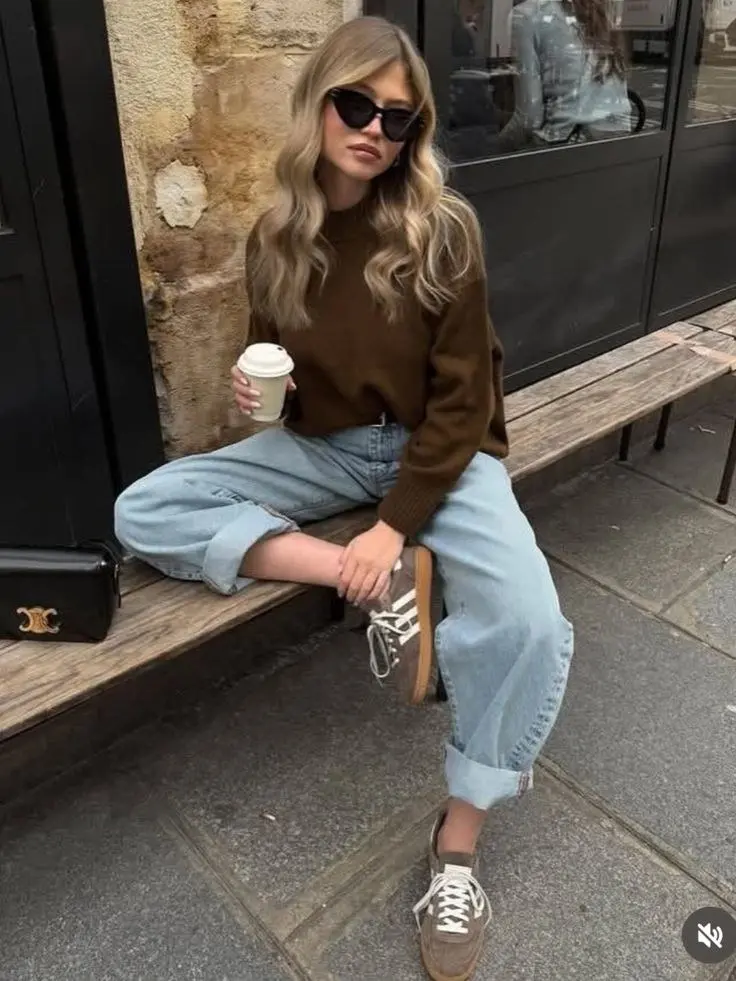 blonde woman in mocha brown mock neck sweater light wash wide leg cuffed jeans taupe gum sole sneakers and small black shoulder bag sitting outside cafe outfit 2
