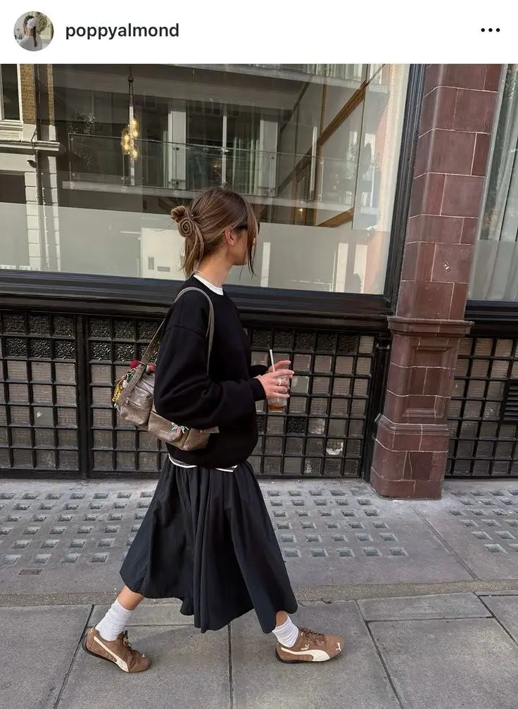 woman in black oversized sweatshirt over white shirt and black pleated midi skirt with brown suede Puma sneakers white ankle socks and brown vintage shoulder bag walking on street outfit 3