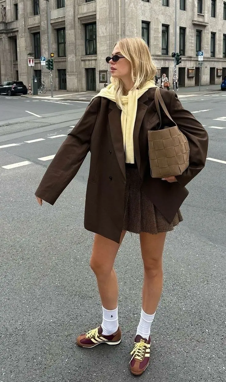 blonde woman in dark brown oversized double-breasted blazer cream yellow hoodie underneath brown plaid pleated mini skirt tan woven tote bag burgundy and yellow stripe sneakers white socks outfit 4