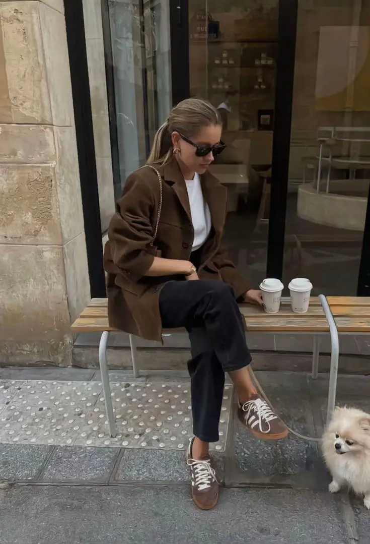 woman in camel brown oversized wool coat white fitted t-shirt black straight leg trousers brown Adidas sneakers gold chain shoulder bag and cat-eye sunglasses sitting outside cafe outfit 5