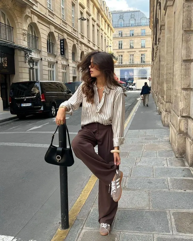 brunette woman in striped cream and tan button-up blouse wide leg dark brown trousers taupe gum-sole sneakers black shoulder bag gold cuff bracelet and round sunglasses standing on street outfit 6