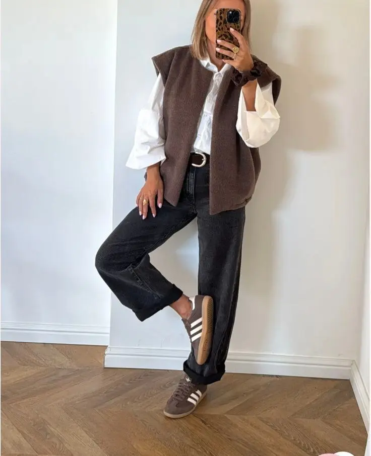 woman in brown knit sweater vest over white puff sleeve shirt dark wash rolled jeans brown leather belt and brown striped Adidas sneakers mirror selfie outfit 7