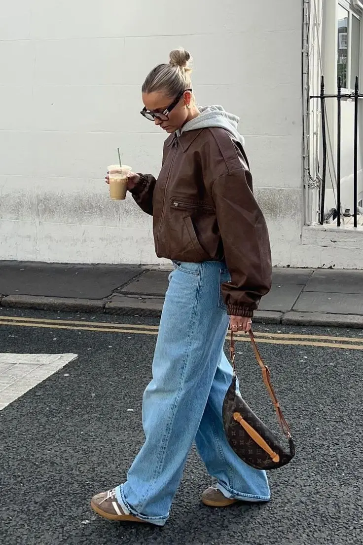 blonde woman in dark brown leather bomber jacket grey hoodie underneath baggy light wash wide leg jeans brown Adidas sneakers and monogram print crescent bag walking on street outfit 8
