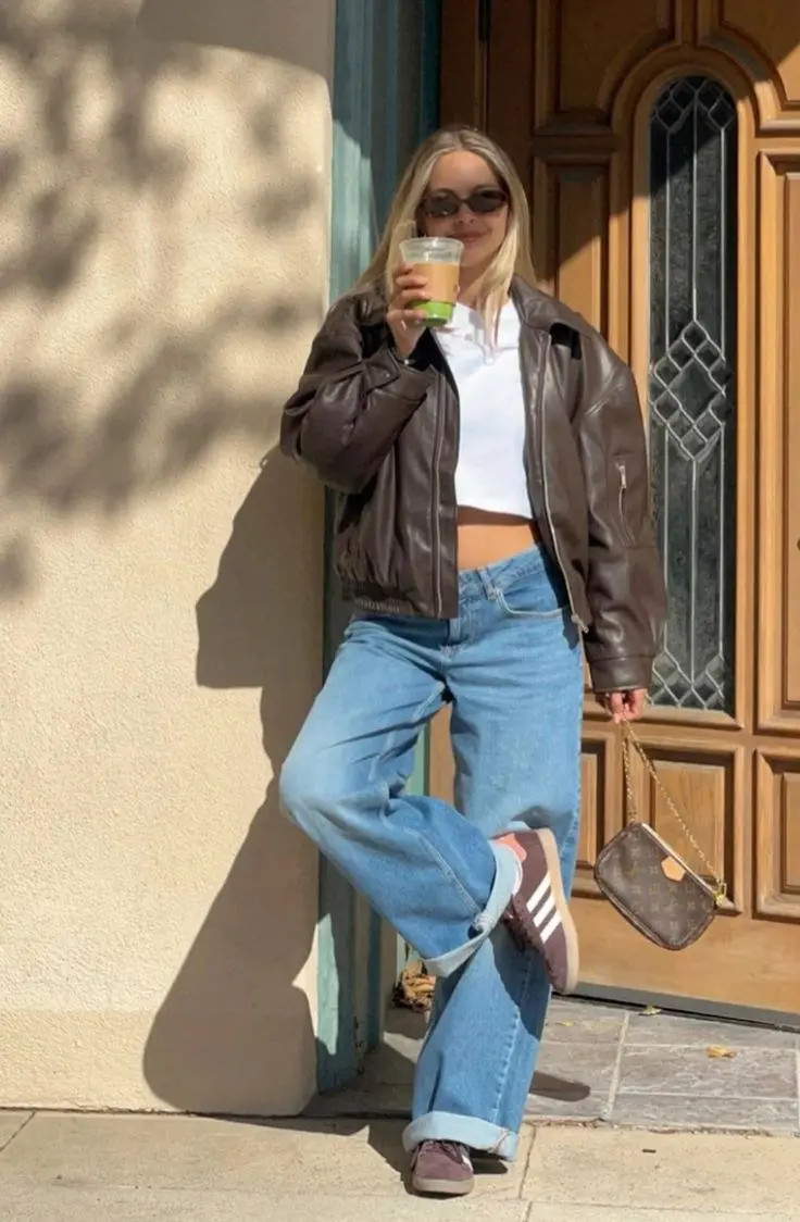 blonde woman in dark brown leather bomber jacket white cropped t-shirt low rise wide leg light wash jeans mauve brown Adidas sneakers small monogram chain bag and oval sunglasses outfit 9