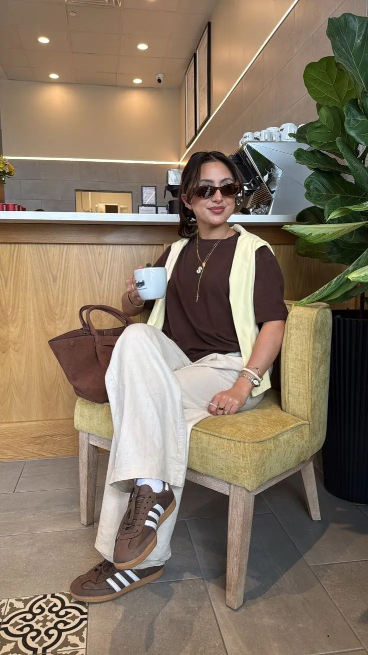 young woman in chocolate brown t-shirt cream wide leg linen pants butter yellow knit vest brown suede Adidas sneakers white socks brown suede tote bag gold layered necklaces and small sunglasses sitting in cafe outfit 10