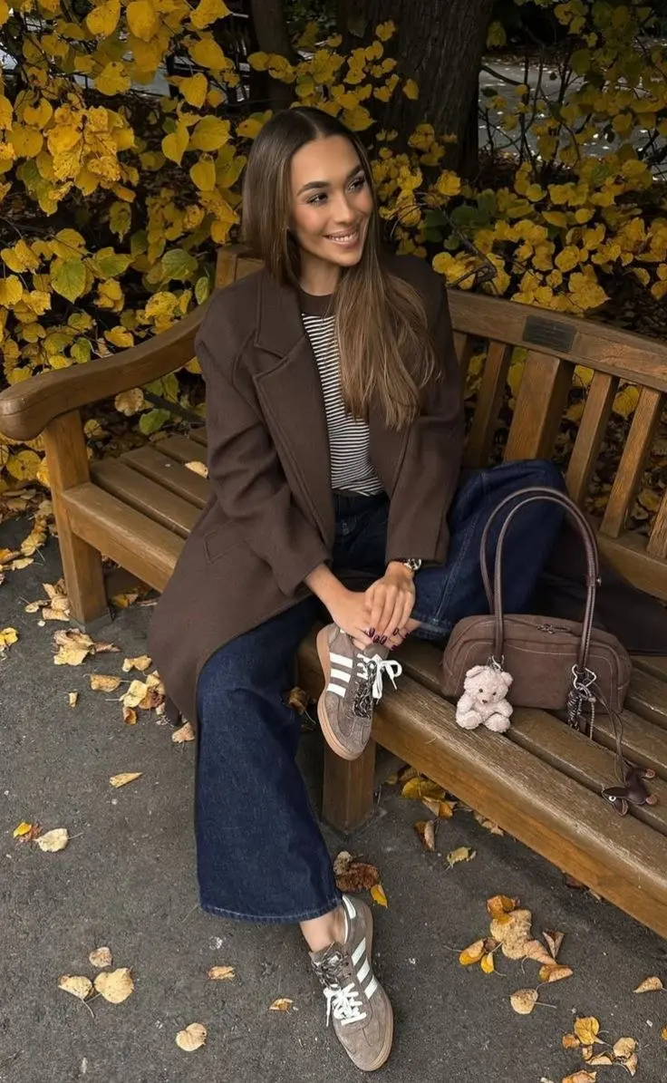 brunette woman in dark brown long wool coat black and white striped t-shirt dark indigo flared jeans taupe Adidas sneakers small brown suede bag with charm sitting on wooden bench with autumn leaves outfit 11