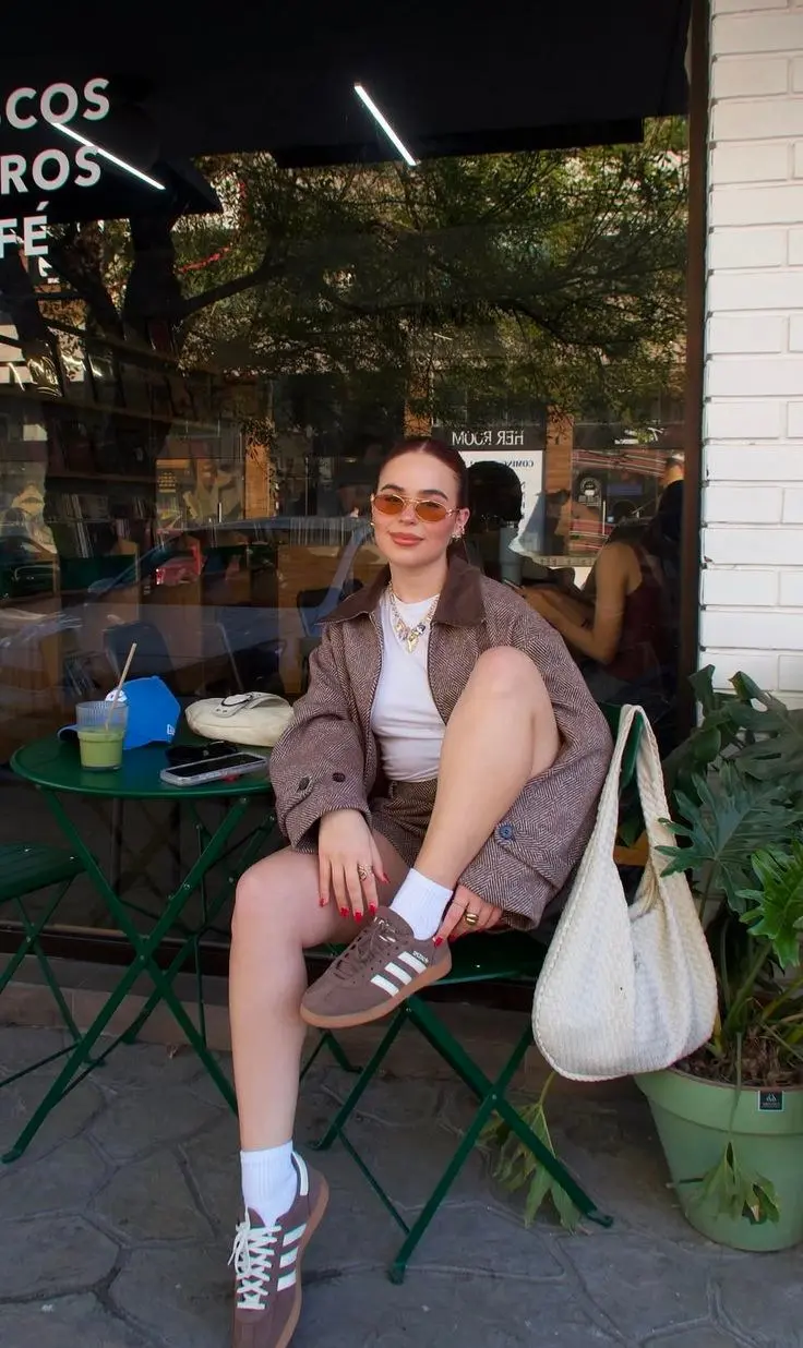 young woman with red nails in brown herringbone oversized jacket white ribbed bodysuit brown herringbone mini skirt brown suede Adidas sneakers white ankle socks cream woven raffia tote bag gold statement necklace small oval sunglasses sitting at outdoor cafe outfit 14