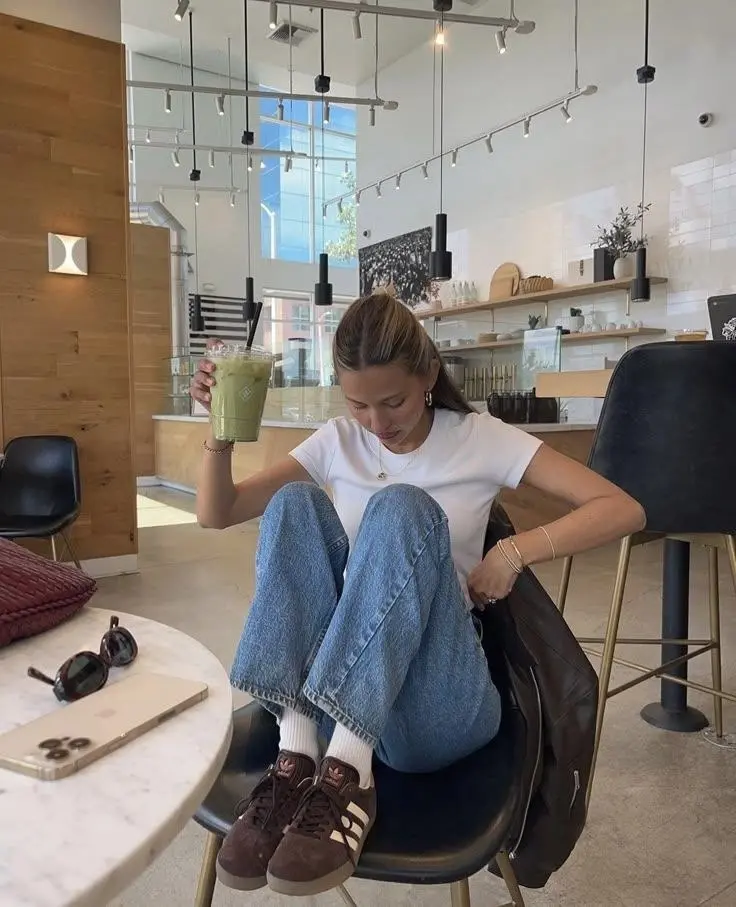 woman in fitted white t-shirt baggy medium wash jeans white ankle socks dark brown Adidas Gazelle sneakers sitting in cafe with sunglasses and gold bracelets outfit 16