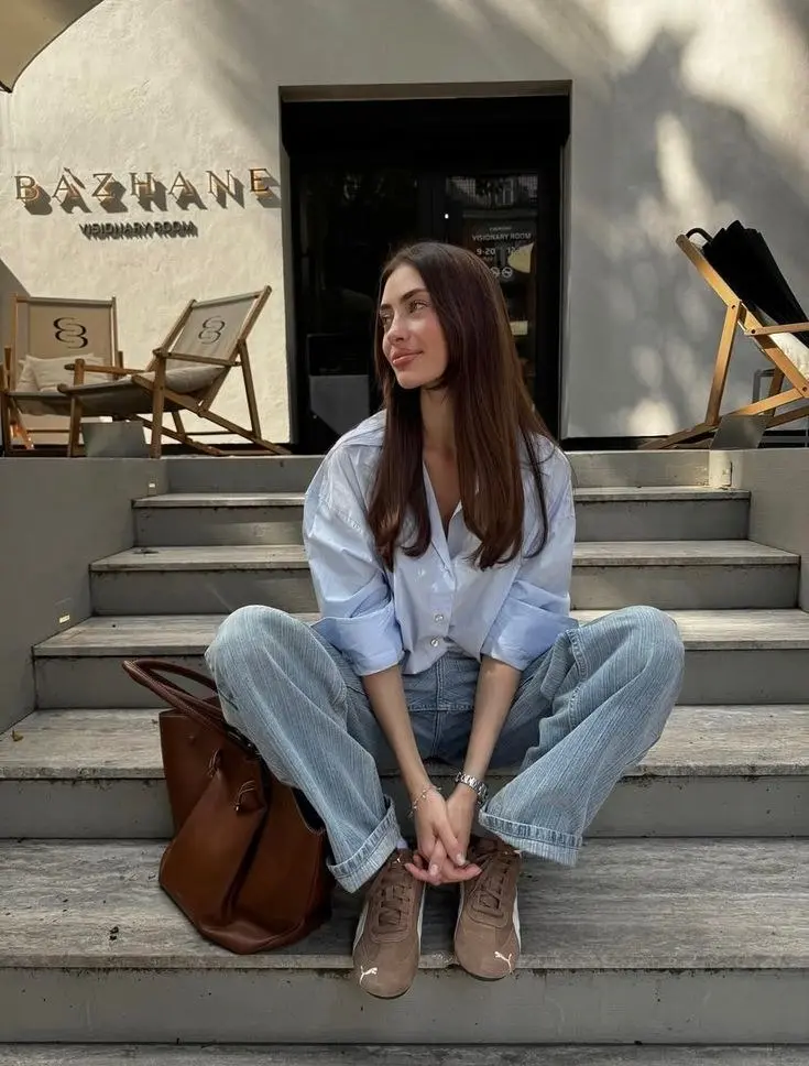 brunette woman in oversized light blue button-up shirt wide leg light wash striped denim jeans tan suede Puma sneakers cognac brown leather tote bag silver bracelet watch sitting on steps outdoor outfit 17
