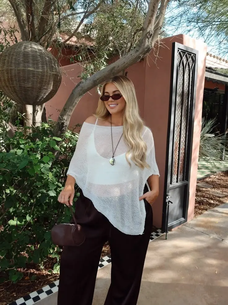 blonde woman in white sheer knit poncho spaghetti strap cami wide black trousers oval sunglasses long pendant necklace and burgundy barrel bag outdoor garden setting outfit 5
