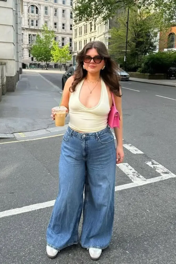 curvy brunette woman in cream deep scoop halter top wide leg mid wash jeans white sneakers pink crossbody bag and large frame sunglasses street outfit 8