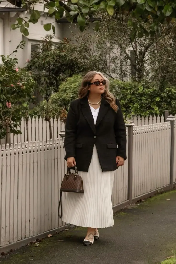 curvy blonde woman in black blazer white top cream pleated midi skirt two tone ballet flats pearl necklace oval sunglasses and structured brown top handle bag outdoors outfit 9