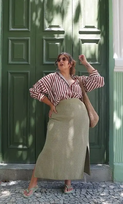curvy woman in burgundy white stripe puff sleeve shirt tucked into sage knit midi skirt green flat sandals woven tote bag and round sunglasses against green door outfit 10