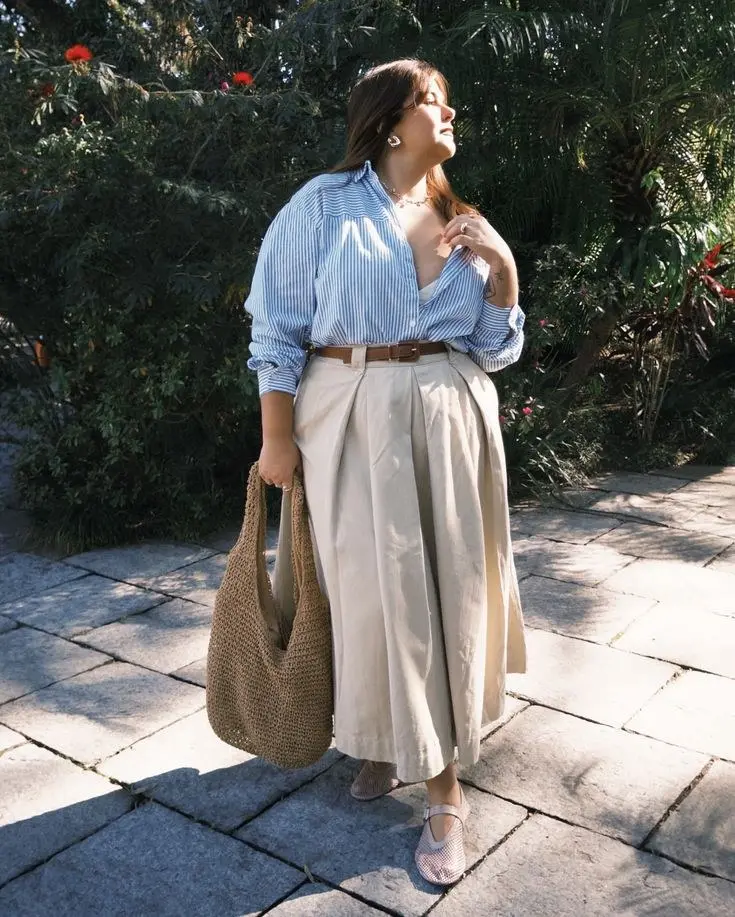 curvy woman in blue white pinstripe oversized shirt belted into wide leg khaki trousers mesh mary jane flats and woven tote bag outdoors garden setting outfit 11