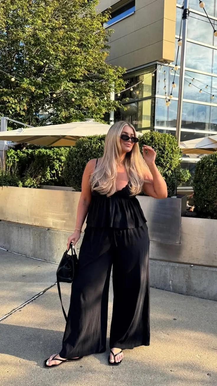 curvy blonde woman in all black ruffle tier cami wide leg satin trousers thong sandals and small black bag outdoor rooftop setting outfit 12