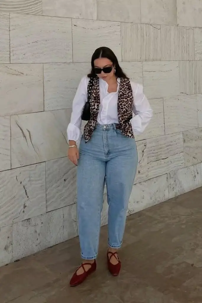 curvy brunette woman in leopard print vest white poplin puff sleeve shirt high rise mom jeans red cross strap flats dark sunglasses and small black bag stone wall outdoor outfit 15