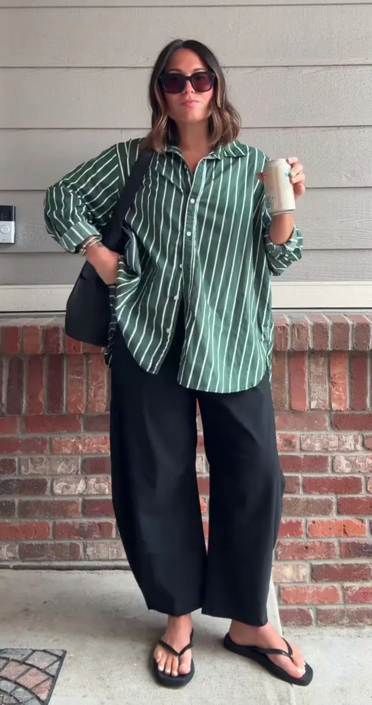 curvy brunette woman in oversized green white stripe button down shirt wide black trousers flat black thong sandals and black crossbody bag outdoors front of house outfit 19
