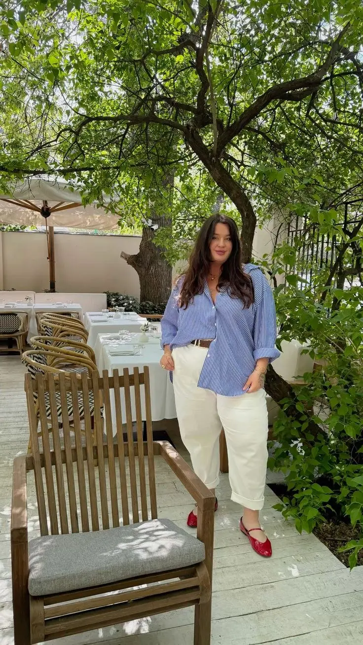 curvy brunette woman in blue white pinstripe oversized shirt tan belt ivory barrel leg cuffed trousers red patent ballet flats and gold necklace outdoor garden restaurant outfit 20