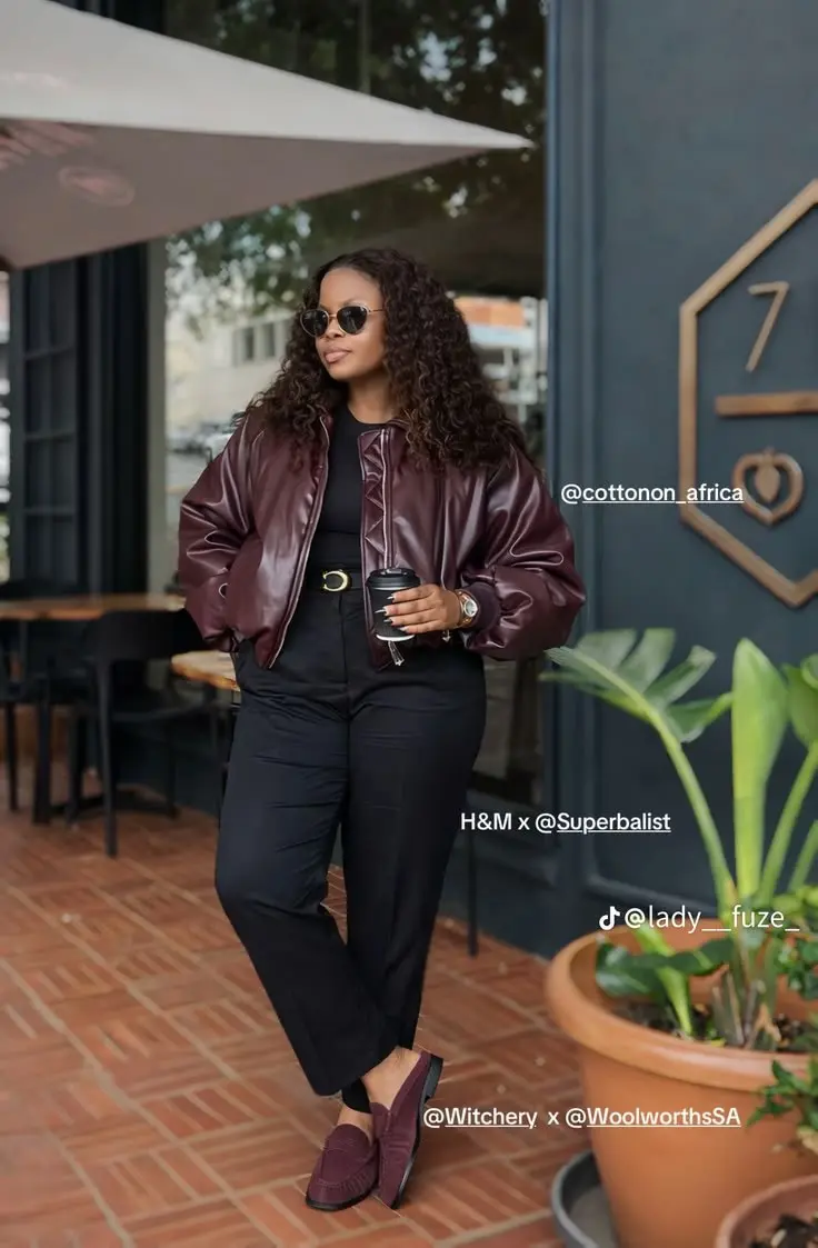 woman in burgundy faux leather bomber jacket black top black straight leg trousers gold belt burgundy loafers round sunglasses holding coffee cup outfit 1