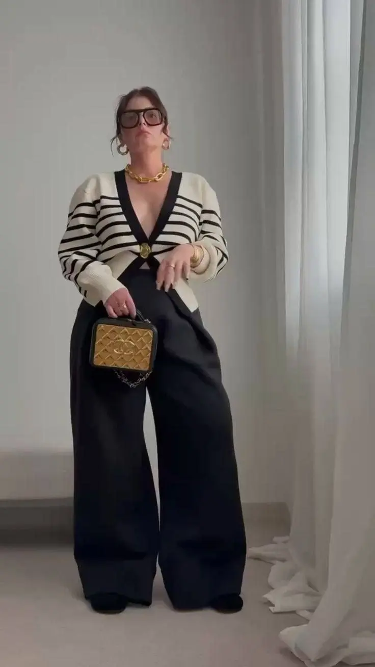 woman in cream black striped deep V oversized cardigan black wide leg trousers chunky gold chain necklace large square glasses gold hoop earrings quilted structured handbag outfit 4