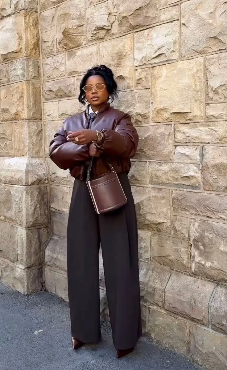 woman in chocolate brown cropped leather bomber jacket white collared shirt dark chocolate wide leg trousers brown bucket crossbody bag pointed brown boots oval gold sunglasses gold jewelry outfit 5