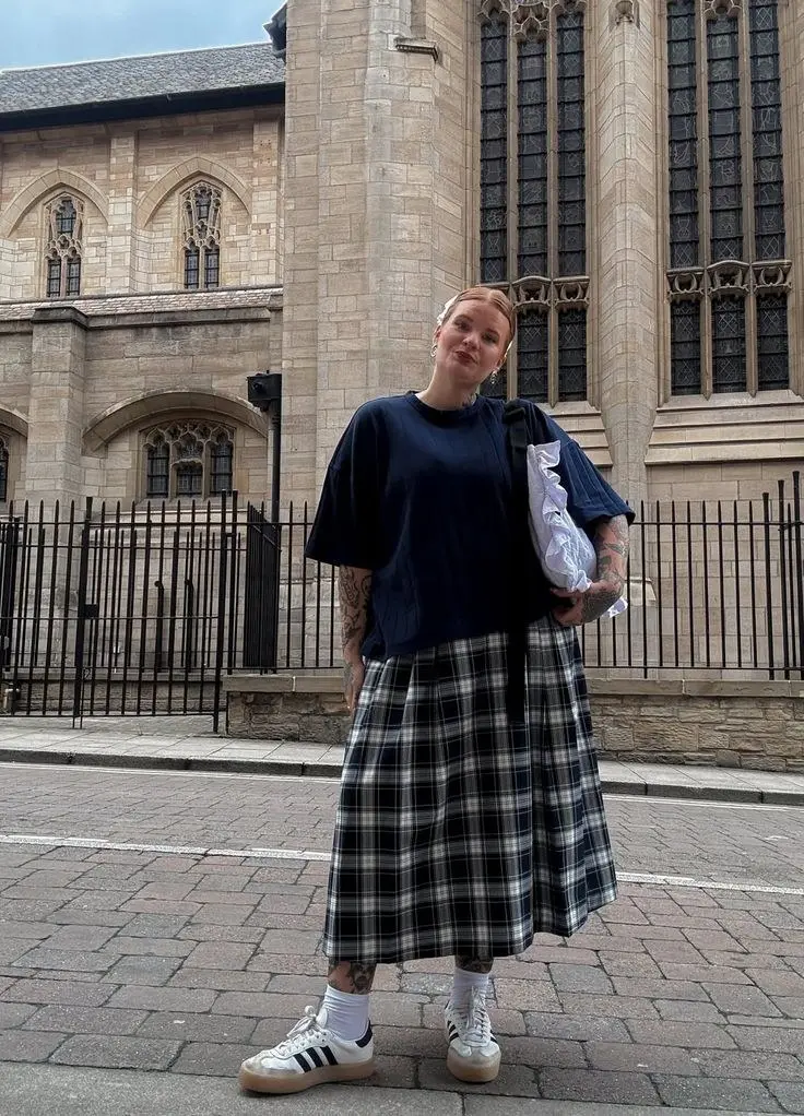 woman in oversized navy blue tee plaid navy white midi pleated skirt white socks adidas samba sneakers silver ear studs stone building background outfit 10
