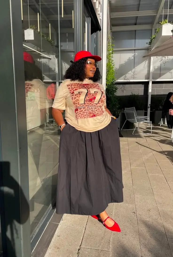 woman in cream graphic print tee dark charcoal voluminous maxi skirt red baseball cap red mary jane flats clear frame glasses outdoor terrace outfit 11