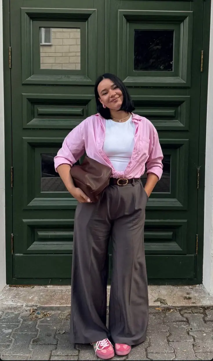 woman in pink oversized open overshirt white fitted tee grey tailored wide leg trousers brown leather belt brown leather clutch bag pink logo sneakers gold necklace earrings green door background outfit 12