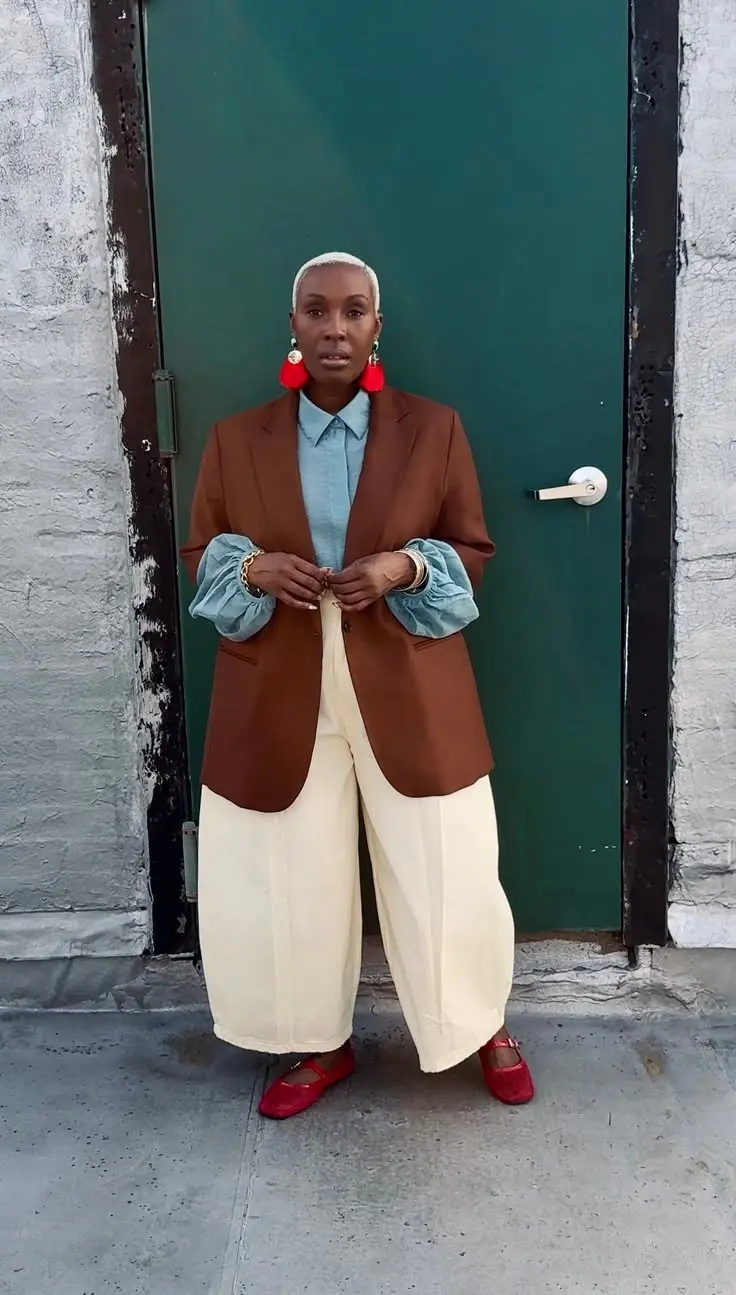 woman in rust brown oversized blazer sky blue puff sleeve shirt cream wide leg trousers red mary jane flats red tassel statement earrings gold bangles white sneaker toe peeking outdoor green door outfit 15