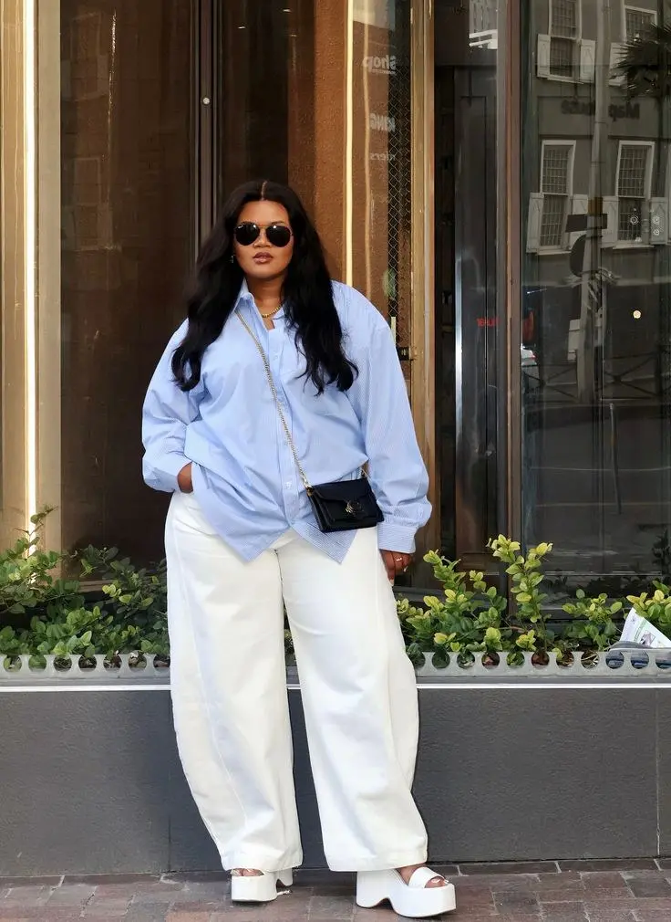 woman in light blue oversized button down shirt white wide leg trousers white chunky platform sandals small black chain crossbody bag gold hoop earrings gold necklace aviator sunglasses outdoor storefront outfit 18
