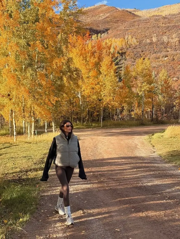 woman in grey cropped puffer vest black shorts sheer tights chunky sneakers walking on autumn trail with orange trees photo 2