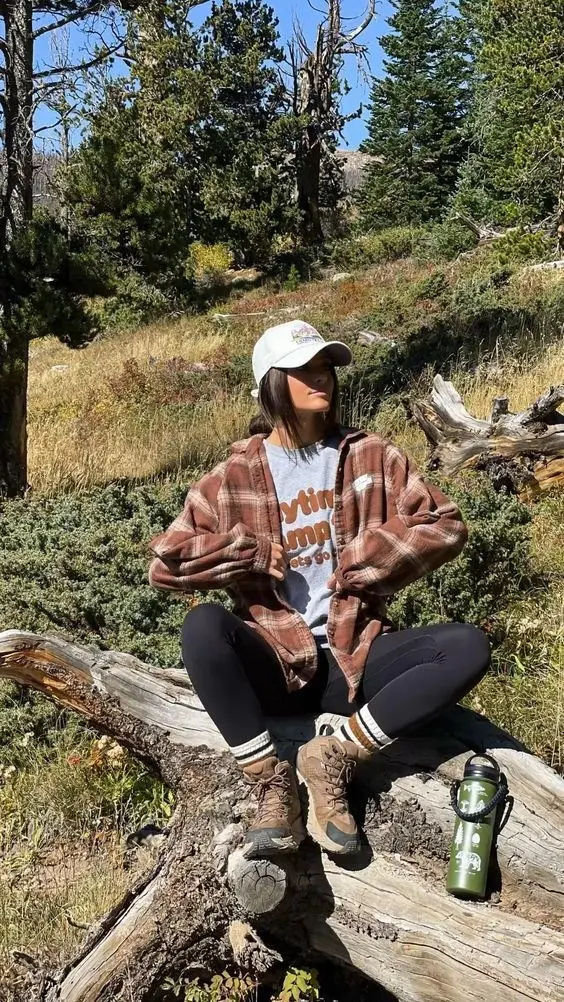 woman in brown plaid flannel shirt grey graphic tee black leggings tan hiking boots white baseball cap sitting on log in mountain forest photo 3