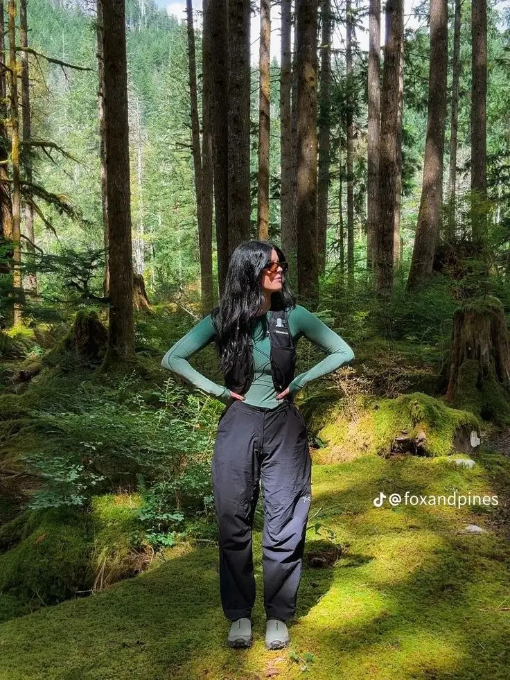 woman in green fitted long sleeve base layer black wide waterproof pants olive trail shoes standing in lush green forest photo 4