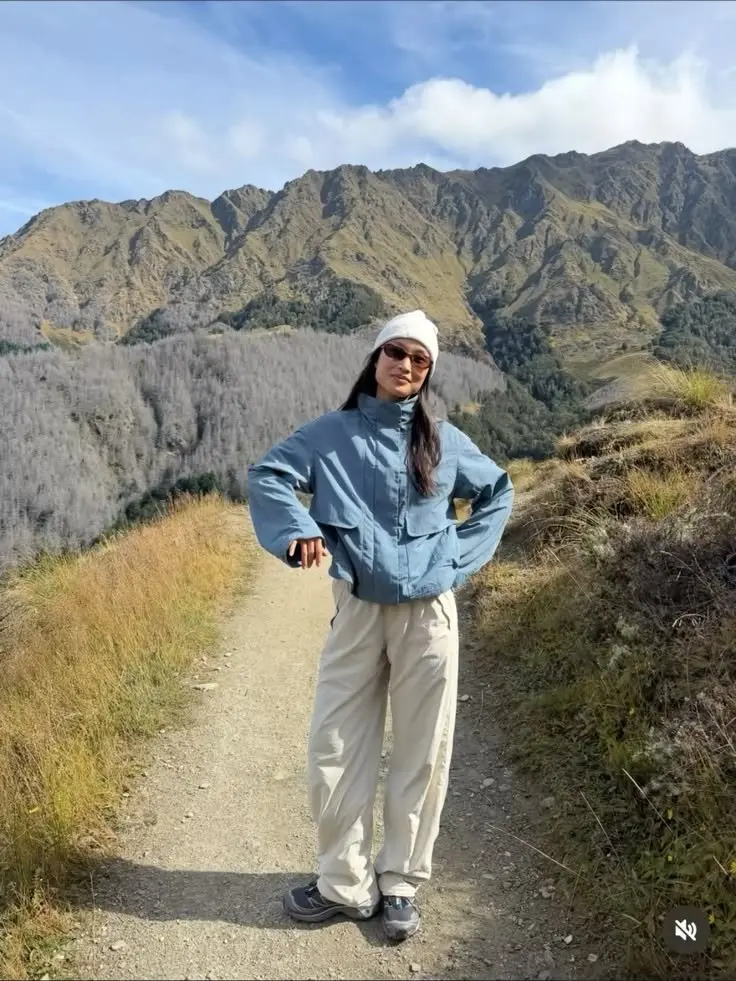 woman in blue shell jacket wide khaki pants grey trail runners white beanie sunglasses on mountain trail photo 5
