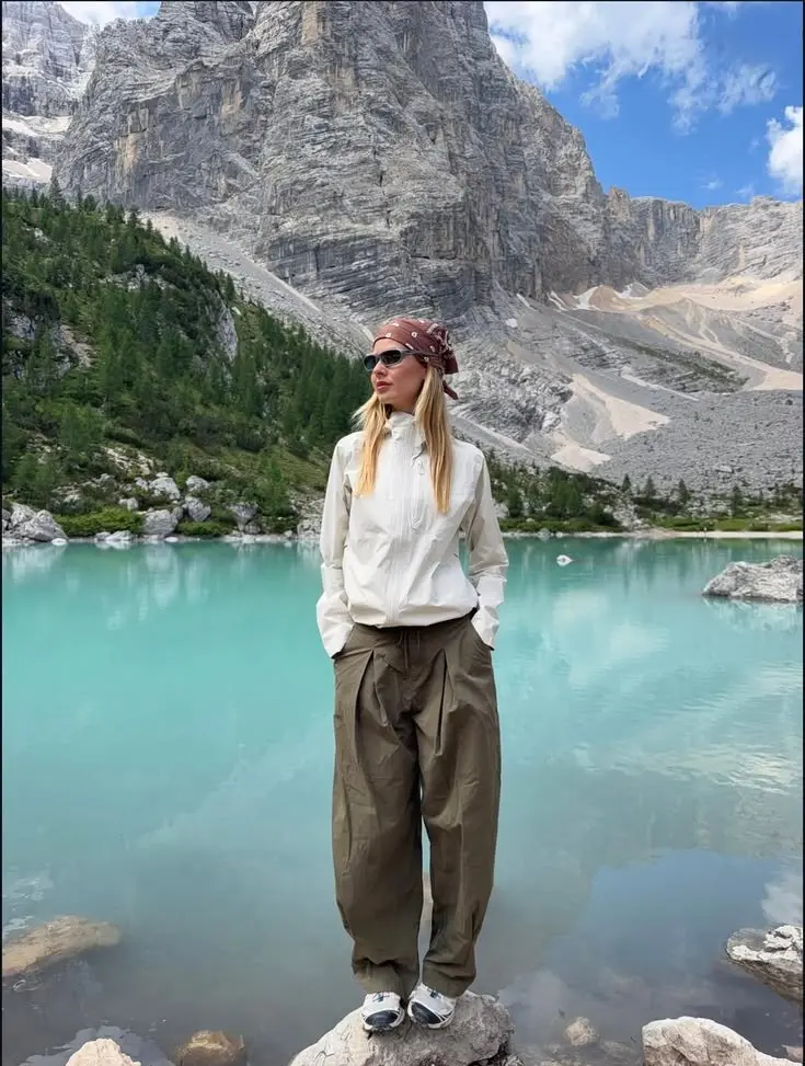 woman in white zip windbreaker wide olive parachute trousers white trail shoes floral bandana headscarf sunglasses standing by turquoise alpine lake photo 6