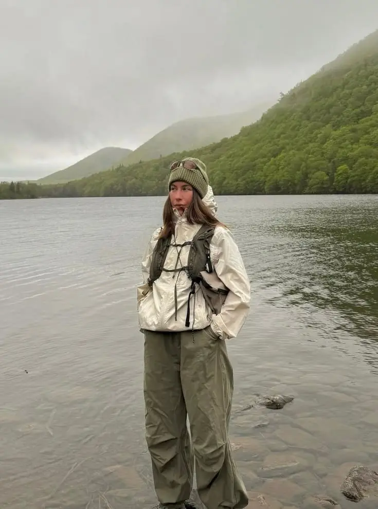 woman in cream packable shell jacket olive wide leg waterproof pants green knit beanie technical backpack sunglasses on beanie standing by misty lake photo 7