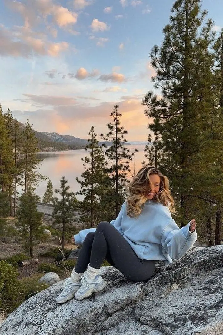 woman in pale blue oversized sweatshirt dark grey leggings white crew socks white chunky sneakers sitting on rocky overlook above lake in pine forest photo 8